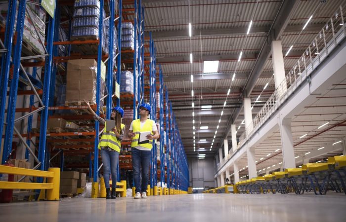 Shot of two workers walking through large warehouse center, observing racks with goods and planing distribution to the market.